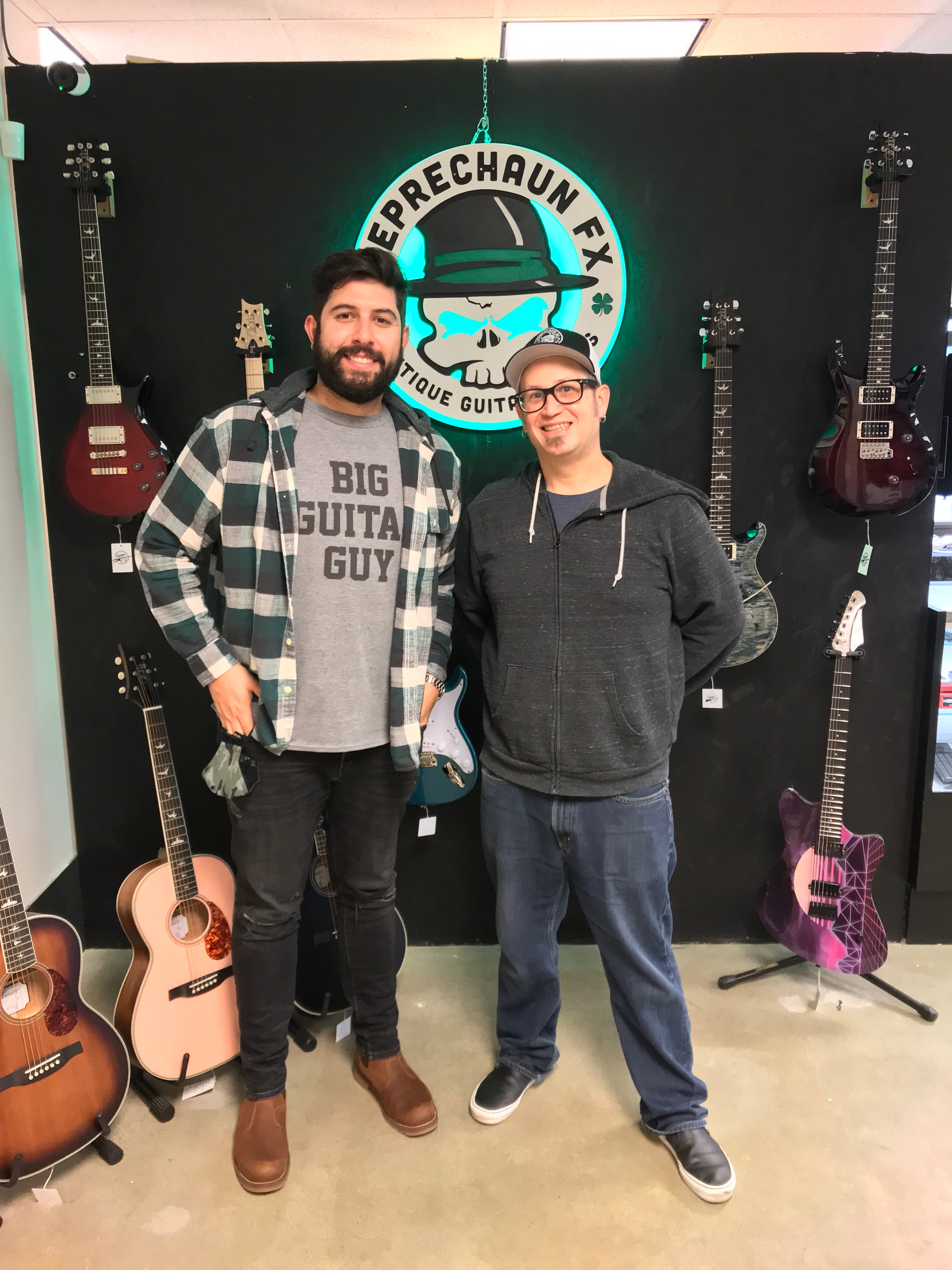 Patrick O'Brien and Brandon Parenteau of Leprechaun FX and Guitar Brando standing infront of a wall of guitars and LED sign of Leprechaun FX